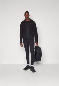 Sort fleece jakke med lynlås, sort t-shirt, sorte leggings og sorte sneakers. Holder en sort rygsæk med netlomme på siden.
