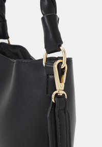 Zbliżenie na rączkę czarnej skórzanej torebki z zapięciem i elementami metalowymi w złotym odcieniu na jednolitym jasnym tle.