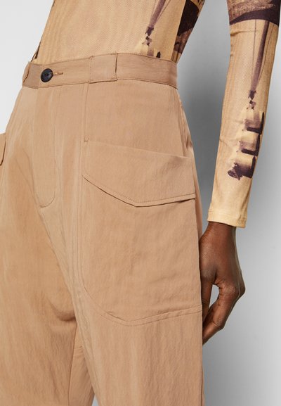 Calças bege feitas de um tecido texturizado, com fecho de botão e bolsos laterais com detalhes de aba. Corte ajustado com um design simples.