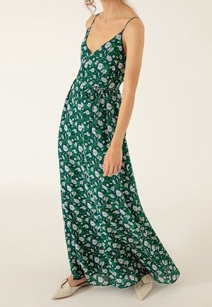 Woman wearing green sleeveless floral maxi dress with spaghetti straps and white pointed flats, standing against a plain background.