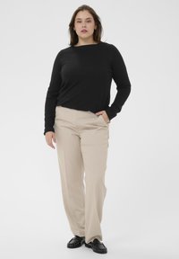 Chemise noire à manches longues associée à un pantalon beige à taille plate. La tenue présente une coupe décontractée et est complétée par des mocassins noirs.