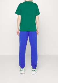 Vihreä puuvillapaita lyhyillä hihoilla, yhdistettynä sinisiin verryttelyhousuihin, joissa on valkoiset sivuraitat. Jalkineina ovat valkoiset lenkkarit, joissa on monivärisiä yksityiskohtia.