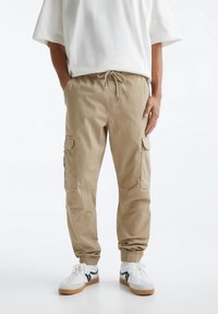 Beige cargo bukser lavet af bomuld, med elastisk talje og snøre, sidelommer og elastiske manchetter ved anklerne.