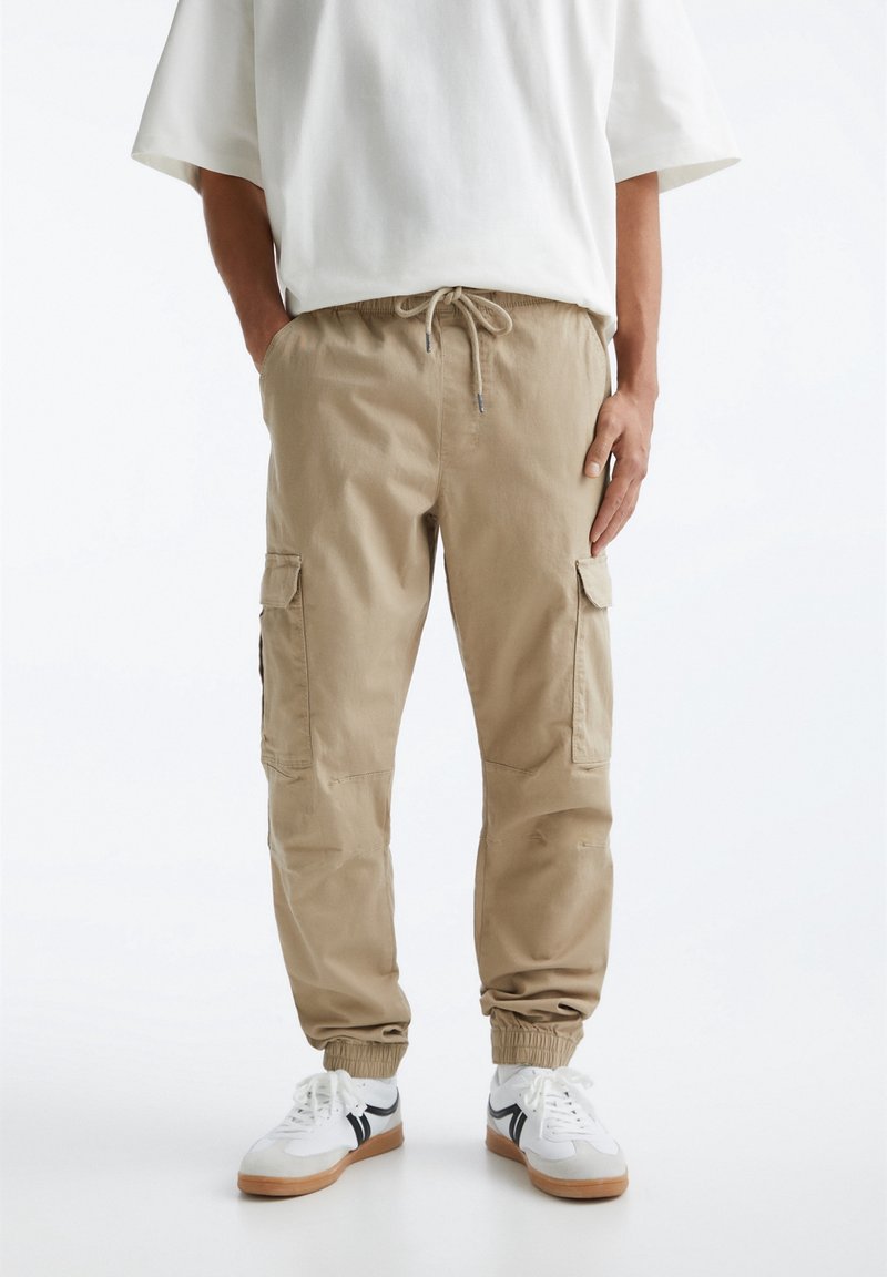 Beige cargo bukser lavet af bomuld, med elastisk talje og snøre, sidelommer og elastiske manchetter ved anklerne.