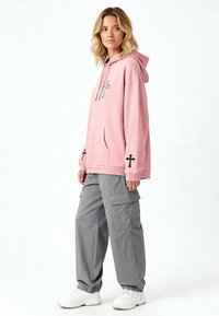 Felpa rosa chiaro con tasca frontale, con design incrociato neri sulle maniche, abbinata a pantaloni cargo grigi e sneakers bianche.