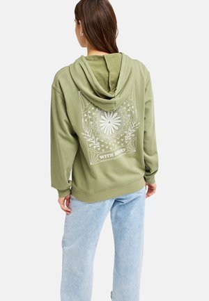 Femme portant un sweat à capuche vert clair avec un motif floral blanc et le texte "WITH HEART" au dos, associée à un jean bleu clair, debout de dos.