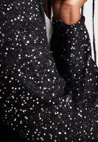 Tissu tricoté orné de sequins noirs, présentant une surface texturée avec de petits sequins réfléchissants répartis uniformément sur le matériau.