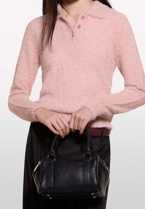 Kvinde iført en pink, bølget sweater og mørke bukser med pindestregsmønster holder en lille sort struktureret lædertaske med dobbelte håndtag.