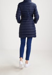 Marineblauwe pufferjas met een horizontaal gestikte ontwerp, hoge kraag en een middellange snit, gedragen met blauwe spijkerbroeken en witte sneakers.