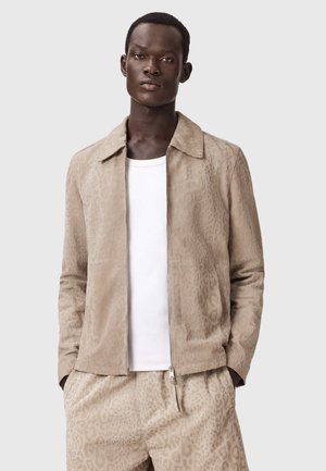 Mann, der eine beige Jacke mit Leopardenmuster und passende Hose über einem weißen Hemd trägt, steht mit den Händen in den Taschen vor einem schlichten Hintergrund.