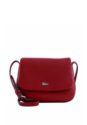 Sac bandoulière en cuir rouge avec fermeture à rabat et petit logo crocodile argenté sur le devant, bandoulière réglable enroulée à côté.