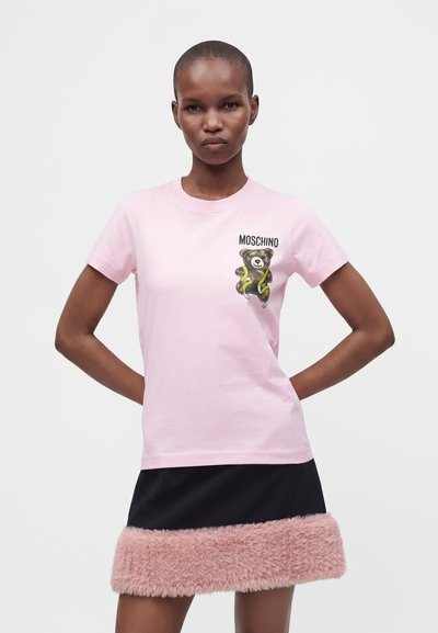 Γυναίκα που φοράει ένα ανοιχτό ροζ t-shirt Moschino με γραφικό αρκουδάκι και μια μαύρη φούστα με ροζ χνουδωτή διακόσμηση, στέκεται με τα χέρια πίσω από την πλάτη.