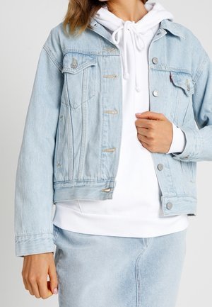 Personne portant une veste en denim bleu clair sur un sweat à capuche blanc et une jupe en denim bleu clair assortie, main ajustant la veste près de la poitrine.