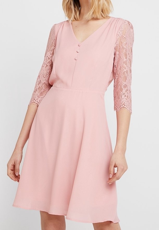 Femme portant une robe rose mi-longue avec des manches trois-quarts en dentelle et trois boutons sur le corsage devant, sur un fond uni.