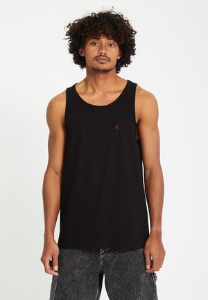 Homme aux cheveux bouclés portant un débardeur noir sans manches et un short en jean gris foncé sur un fond blanc.