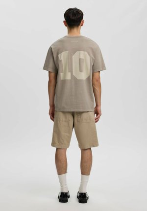 Homme portant un short beige et un t-shirt taupe avec un grand numéro 10 dans le dos, debout, dos à un fond clair uni.