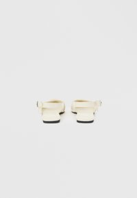 Επίπεδα σανδάλια σε εκρού απόχρωση με λουράκια στον αστράγαλο και μαύρες σόλες, εμφανιζόμενα από πίσω σε απλό ανοιχτό φόντο.