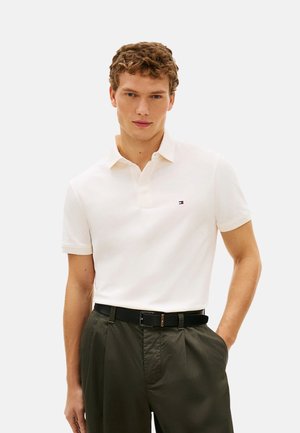 Joven con camiseta polo blanca con un pequeño logo y pantalones verde oscuro, de pie con una mano en el bolsillo frente a un fondo liso.