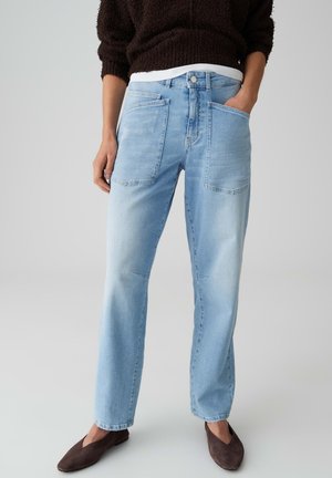 Persona che indossa jeans di denim azzurro chiaro con grandi tasche frontali, maglione marrone scuro peloso e ballerine marroni con punta appuntita, in piedi contro uno sfondo semplice.