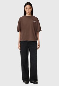 Camiseta grande de color marrón con cuello redondo y mangas cortas; presenta el texto blanco "ALLSAINTS" en la parte delantera, combinada con jeans de pierna ancha negros.