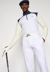 Camisa de golfe de mangas compridas branca com detalhes em azul-marinho, combinada com calças brancas e um cinto branco. O modelo segura um taco de golfe e usa um boné e uma luva.