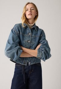 Jaqueta de denim azul clara com um corte cropped, mangas grandes e volumosas, e fecho de cinco botões, combinada com calças largas de denim azul escuro.