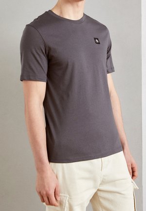 Homme portant un t-shirt gris foncé ajusté à manches courtes avec une petite écusson noir sur la poitrine et un pantalon cargo beige clair.