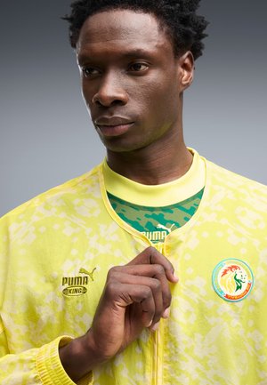 Hombre con una chaqueta amarilla Puma King con el logo de la Federación de Fútbol de Senegal, sosteniendo la cremallera de la chaqueta cerca del cuello.