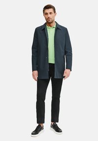 Granatowa kurtka zewnętrzna, zielona koszulka polo, czarne spodnie i czarne sneakersy. Kurtka ma klasyczny kołnierzyk i zapięcie na guziki, gładką teksturę.