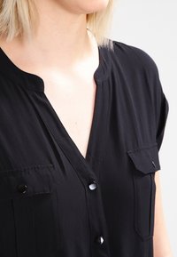 Blouse noire à manches courtes avec un col en V, deux poches poitrine, accents de boutons noirs et une texture de tissu lisse.