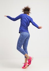 Niebieska bluzka z długim rękawem i niebieskie legginsy w wzory. Różowe buty sportowe z czarnymi detalami. Osoba uchwycona w dynamicznej pozie.
