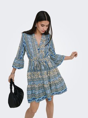 Femme portant une robe bleue à motifs longueur genoux avec manches 3/4, tenant un sac à main noir froncé dans sa main gauche, regardant vers le bas.