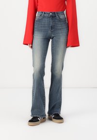 Blå flared jeans med en falmet design, højt liv og fem lommer, parret med sorte sneakers med hvide og grå striber.