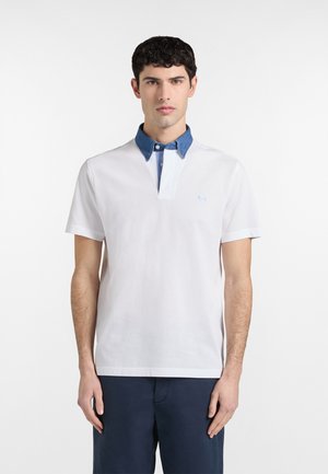 Mężczyzna ubrany w białą, krótkorękawową koszulkę polo z niebieskim, dżinsowym kołnierzem i ciemnymi spodniami, stojący na tle jednolitego, białego tła.