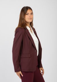 Blazer su misura bordeaux con spalle definite, chiusura con un solo bottone frontale e due tasche laterali. Indossato sopra una camicetta bianca.