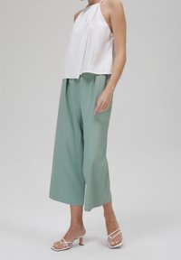 Haut blanc à col licou, associé à un pantalon ample vert clair à jambes larges. Les sandales ont un talon bloc et de fines lanières blanches.