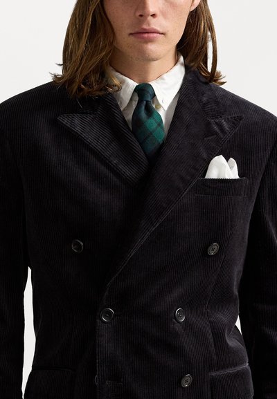 Man die een zwart corduroy dubbelbreasted blazer, een wit shirt, een groen en zwart geruit stropdas, en een witte pochet draagt.