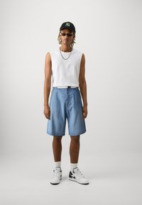 Fehér ujjatlan póló, világoskék farmer rövidnadrág, fekete logós sapka, ezüstlánc és fehér sportcipő fekete kiegészítőkkel. Egyszerű háttér.