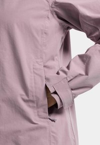 Person wearing a long-sleeved lavender shirt with their right hand placed inside a side pocket, set against a plain background.