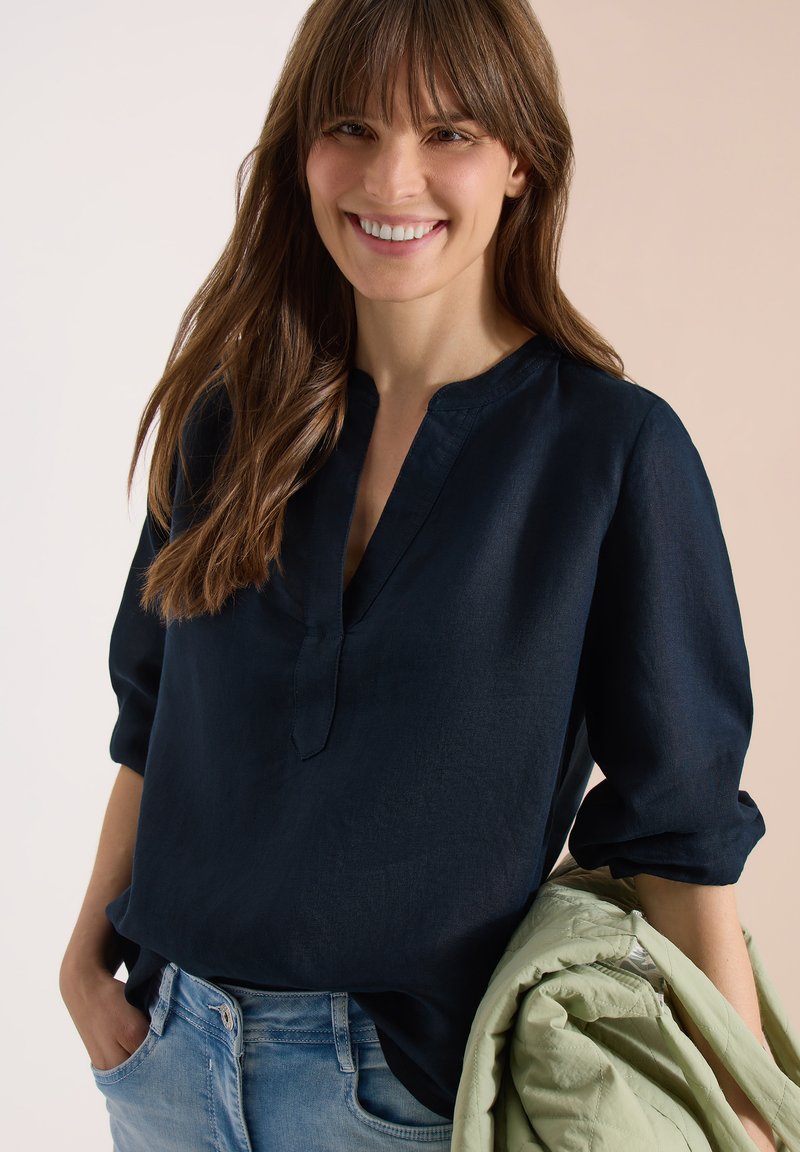 Femme souriante, portant un chemisier bleu marine à col en V avec manches retroussées et un jean bleu clair, tenant une veste vert clair dans son bras gauche.