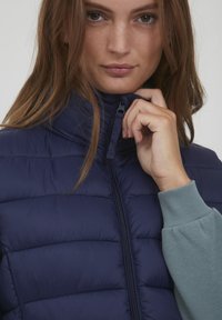 Marineblauwe gewatteerde vest met hoge kraag, ritssluiting en kanalenstiksels. Model draagt er een lichtblauwe trui onder. Soepele textuur, casual pasvorm.