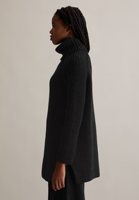 Schwarzer, strukturierter Rollkragenpullover mit lockerer Passform, seitlichen Schlitzen und geripptem Kragen. Der Stoff zeigt ein markantes gewebtes Muster.