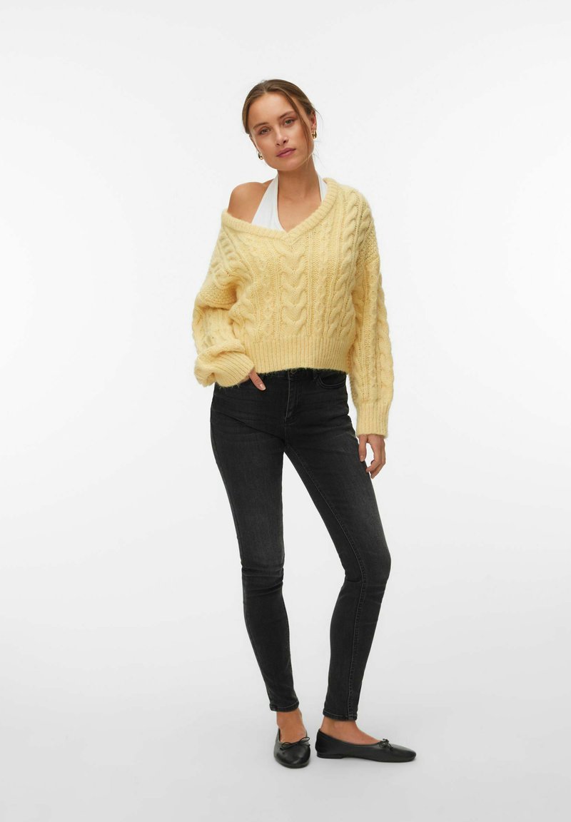 Femme portant un pull jaune ample en maille torsadée par-dessus un haut blanc, un jean skinny noir et des ballerines noires, debout devant un fond uni.