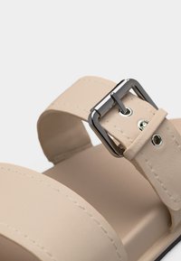 Μπεζ λουρί σανδαλιού με ασημένια μεταλλική αγκράφα και ραφή σε φόντο από λείο πέλμα.