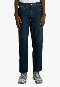 Jeans in denim blu scuro con design a gamba dritta, chiusura con bottone in metallo e cuciture a contrasto. Indossati con scarpe sportive grigie.