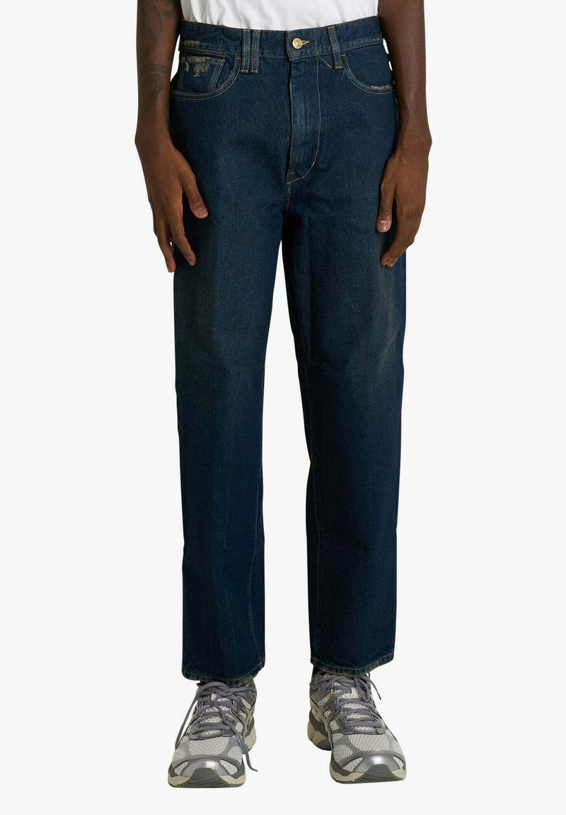 Jeans in denim blu scuro con design a gamba dritta, chiusura con bottone in metallo e cuciture a contrasto. Indossati con scarpe sportive grigie.