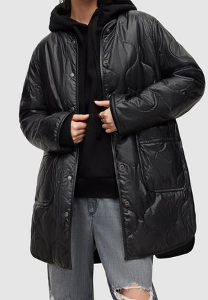 Un mannequin porte un manteau noir matelassé par-dessus un sweat à capuche et un jean gris déchiré, et tient les pans du manteau devant un fond uni.
