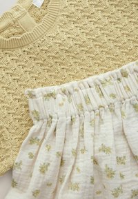 Stickad gul topp med texturerat mönster, tillsammans med en vit kjol med grönt blommönster och ett elastiskt midjeband.