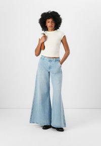 Lyseblå slengbukser med høy midje, laget av denim. Kombinert med en tettsittende kremfarget topp. Modellen står med hendene på hoftene.
