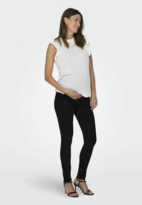 Haut blanc texturé avec des manches volantées, associé à un jean skinny noir et des talons hauts noirs à lanières. Le mannequin se tient avec une main sur son ventre.